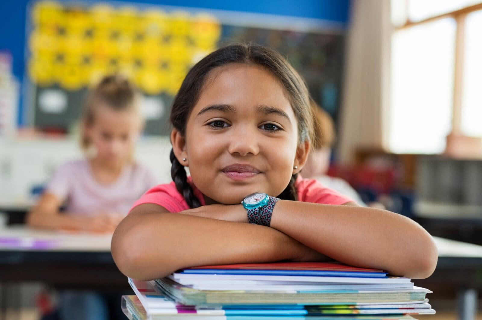 Elementary aged student with arms crossed on top of books in classroom. 